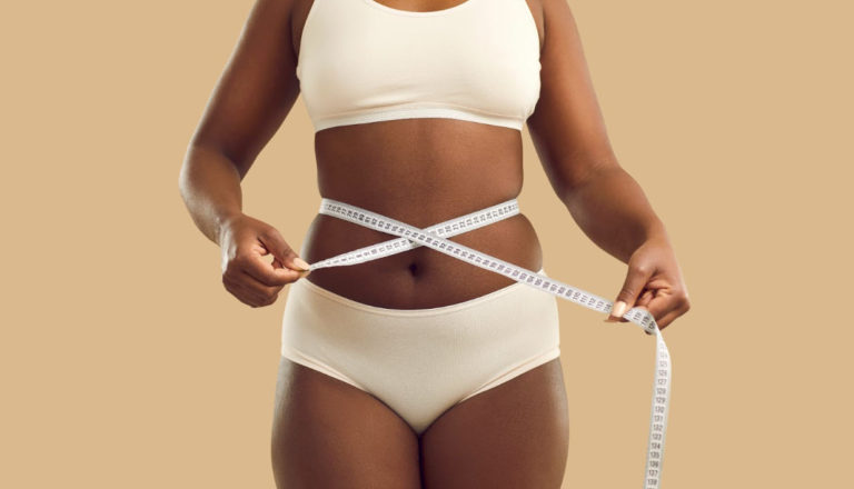 A woman in beige undergarments measuring her waist with a tape measure, symbolizing weight loss progress and the effectiveness of tirzepatide and semaglutide in medical weight management.