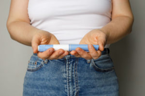 A woman wearing a white tank top and jeans holds a blue and white injection pen in both hands, representing semaglutide as a treatment for weight loss and blood sugar management.