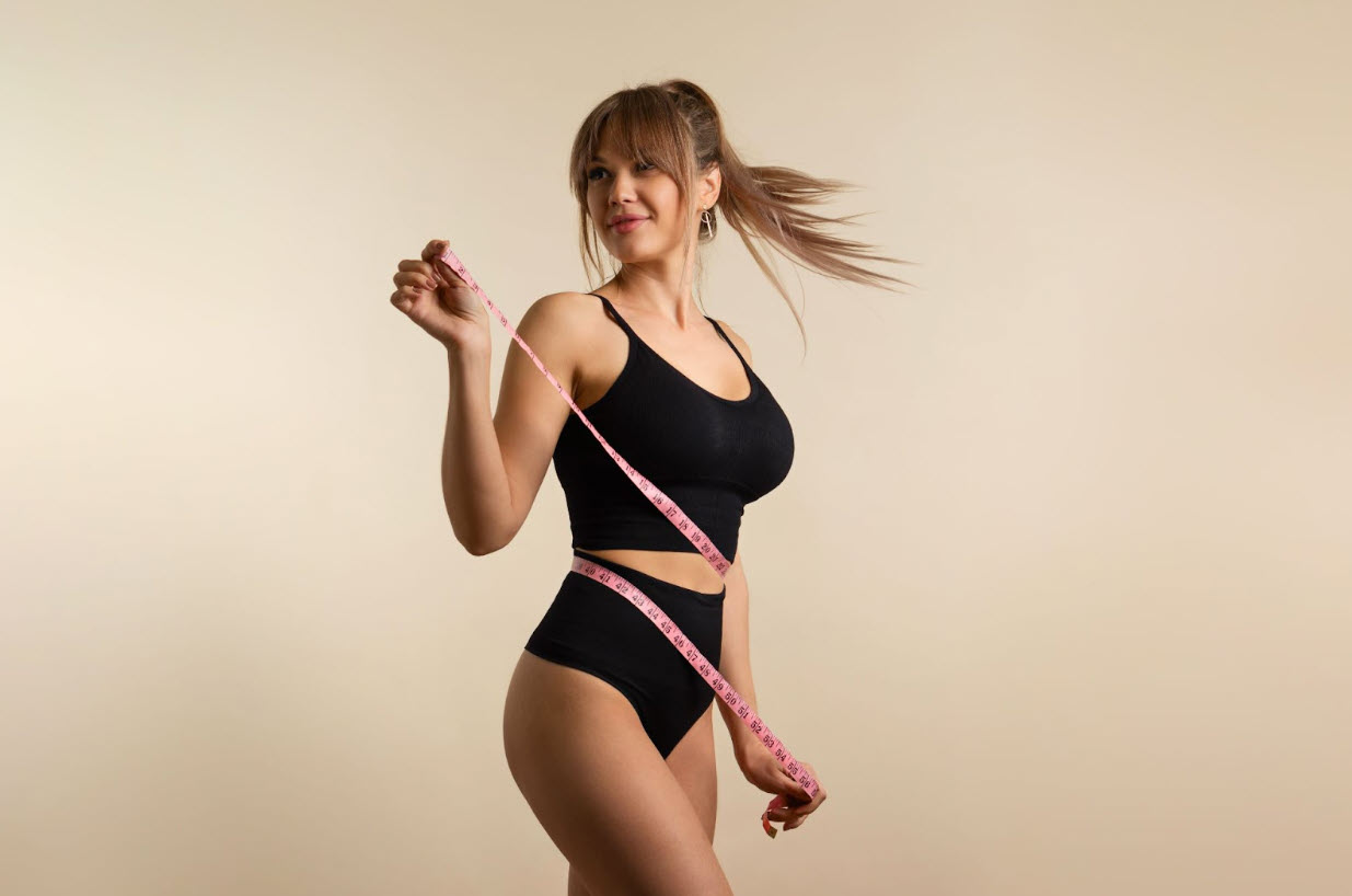 A fit woman in black athletic wear measuring her waist with a pink tape measure, smiling confidently, illustrating the question: does tirzepatide suppress your appetite by helping with portion control and weight management.