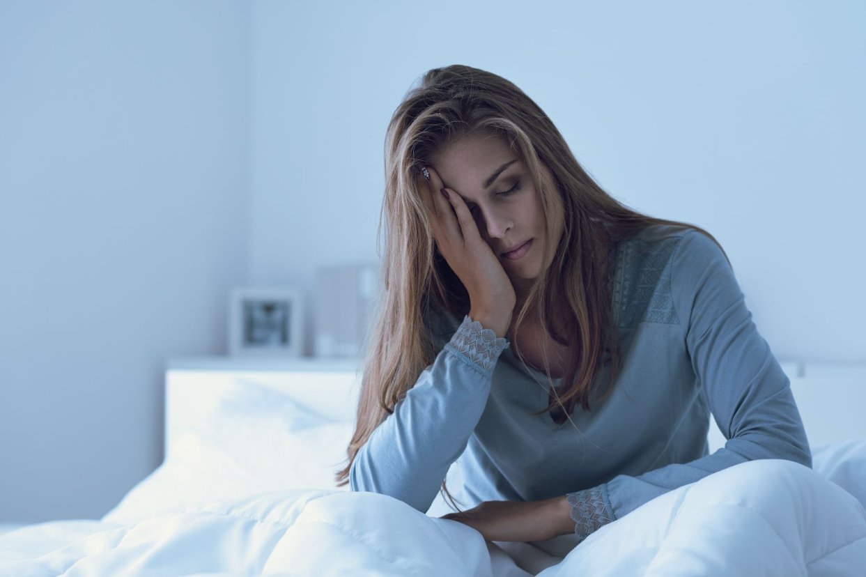 A fatigued woman sits on her bed, holding her head in exhaustion, illustrating the symptoms of hormone imbalance and the benefits of understanding what is bioidentical hormone replacement for restoring energy and well-being.
