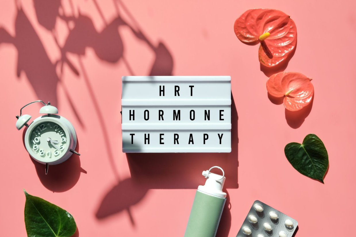 A lightbox with the words 'HRT Hormone Therapy' sits on a pink background alongside a clock, pill blister pack, and skincare bottle, representing the concept of hormone therapy cost and available treatment options.