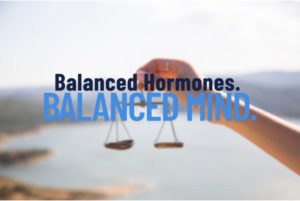 A hand holding a set of scales with the background featuring a blurred view of a lake and hills. The text 'Balanced Hormones. BALANCED MIND.' is prominently displayed over the image, with the words 'Balanced Hormones' in dark blue and 'BALANCED MIND' in a lighter blue.