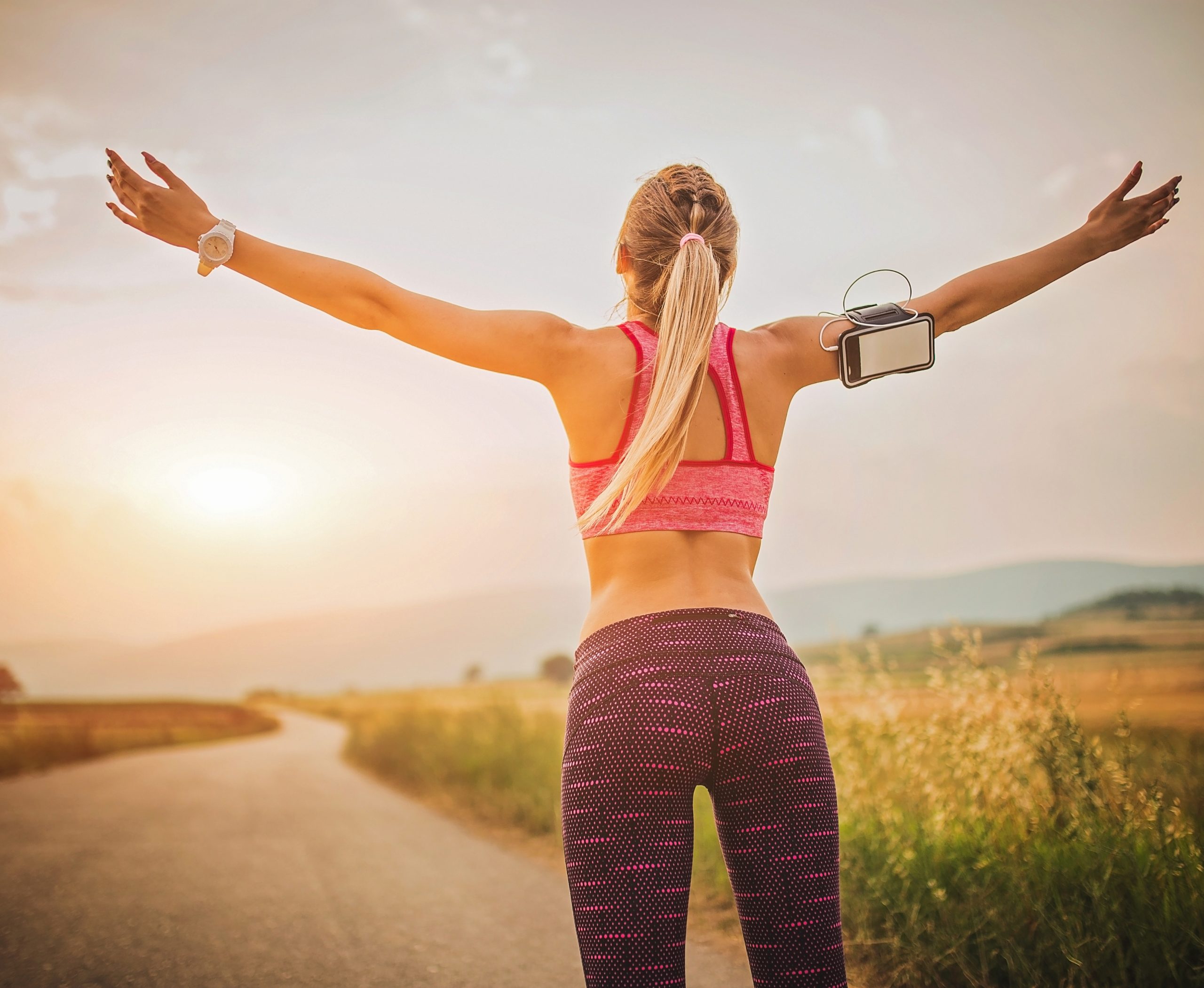 Move. Nourish. Glow. A woman in athletic wear with her arms outstretched, standing on an open road with a scenic landscape in the background. She is wearing a pink sports bra and purple leggings, with a phone armband, as she enjoys the fresh air during her outdoor workout.