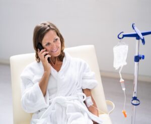 Woman in a white robe receiving IV hydration therapy while smiling and talking on the phone.
